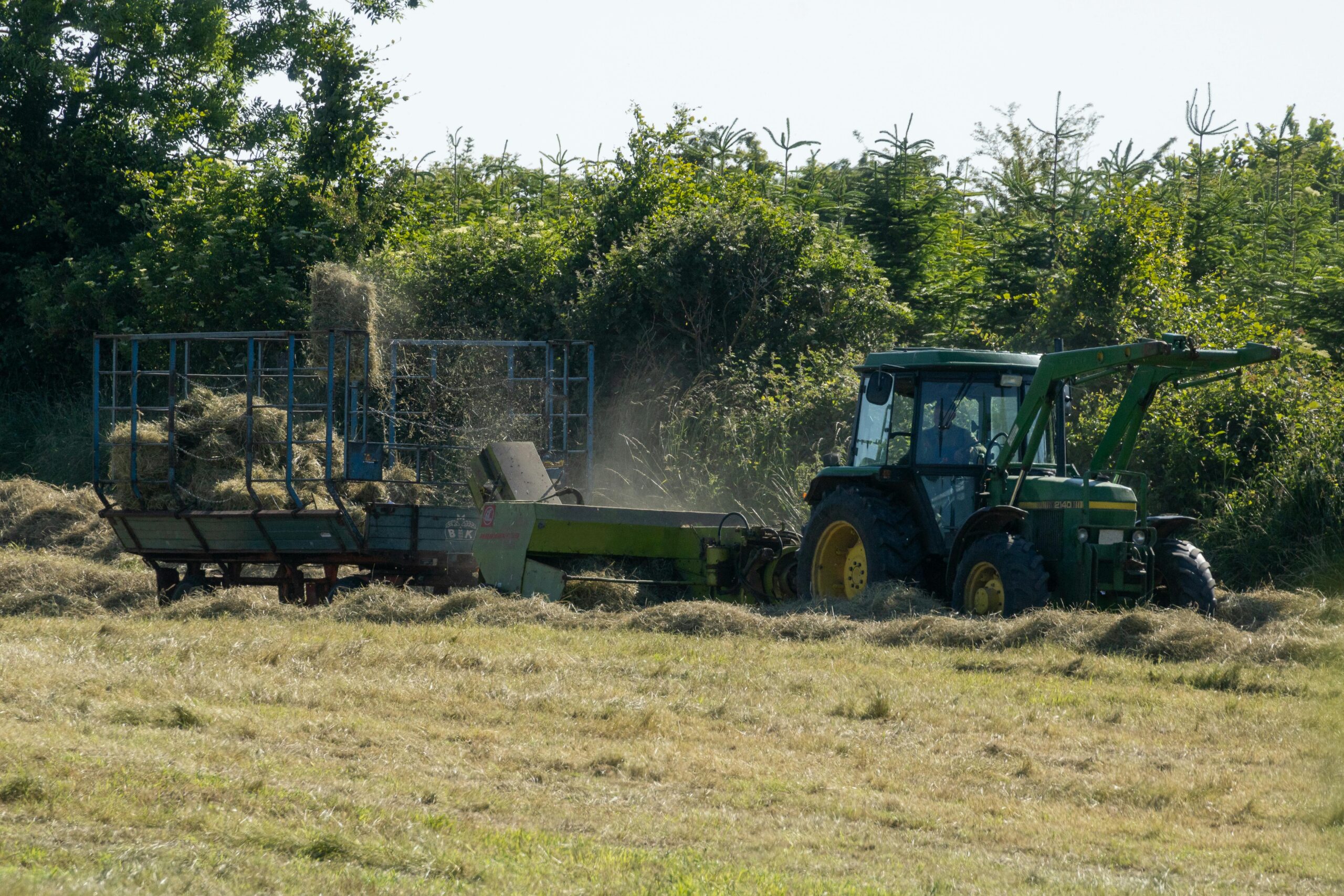A green tractor baling hay in a sunny countryside field, surrounded by greenery.